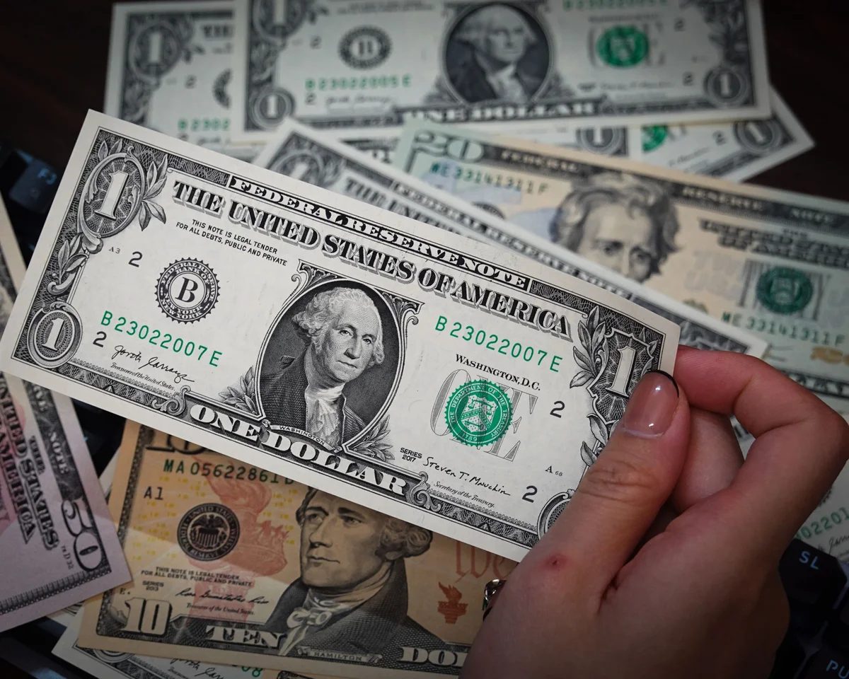 A hand holding a one dollar bill with more currency nearby